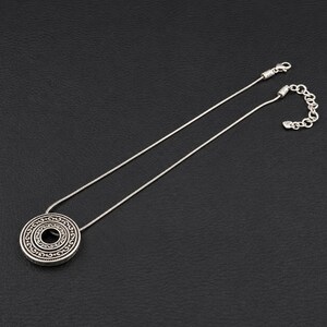 Puede incluir: Un collar de tono plateado con un colgante circular que presenta intrincados patrones de volutas en relieve. El colgante cuelga de una cadena fina y flexible que incluye un cierre de mosquet&oacute;n y una cadena extensora ajustable.