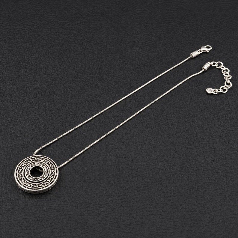 Puede incluir: Un collar de tono plateado con un colgante circular que presenta intrincados patrones de volutas en relieve. El colgante cuelga de una cadena fina y flexible que incluye un cierre de mosquet&oacute;n y una cadena extensora ajustable.