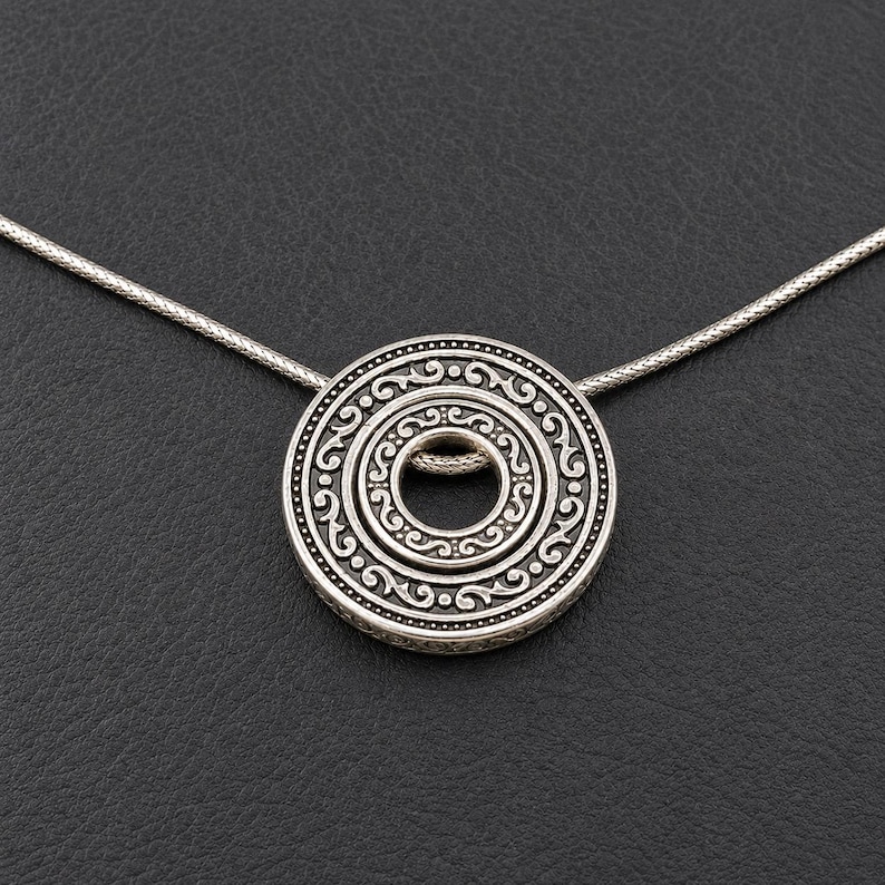 Puede incluir: Un collar con colgante circular de tono plateado que presenta un intrincado patr&oacute;n de volutas en relieve. El colgante consta de dos anillos conc&eacute;ntricos con un recorte circular central, suspendido de una fina cadena de serpiente a juego.