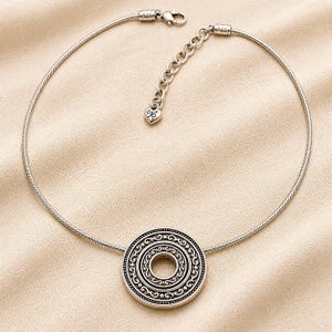 Puede incluir: Un collar de tono plateado con un colgante redondo y texturizado con patrones circulares conc&eacute;ntricos. La cadena es un cord&oacute;n flexible tipo serpiente con cierre de mosquet&oacute;n, una cadena extensora ajustable y un peque&ntilde;o dije en forma de coraz&oacute;n marcado 925.