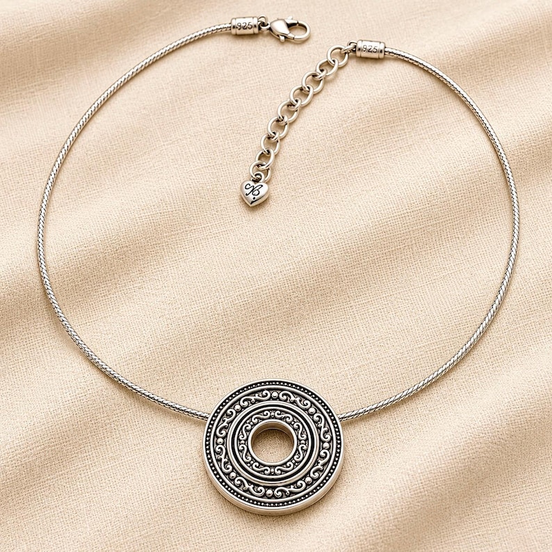 Puede incluir: Un collar de tono plateado con un colgante redondo y texturizado con patrones circulares conc&eacute;ntricos. La cadena es un cord&oacute;n flexible tipo serpiente con cierre de mosquet&oacute;n, una cadena extensora ajustable y un peque&ntilde;o dije en forma de coraz&oacute;n marcado 925.