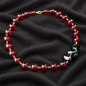 Puede incluir: Un collar que presenta una serie de cuentas redondas y translúcidas de color rojo intenso unidas por un fino hilo de metal dorado. Una cuenta negra única, lisa y asimétrica, se sitúa en el lado derecho, con un cierre de mosquetón dorado.