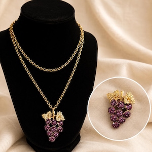 May include: A gold-toned necklace featuring a pendant shaped like a bunch of grapes. The grapes are made of small, round, purple gemstones. The necklace is displayed on a black mannequin, with a close-up of the pendant in a circle.