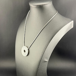 Puede incluir: Un collar con colgante circular de tono plateado que presenta un intrincado patr&oacute;n geom&eacute;trico repetitivo grabado en su superficie. El colgante est&aacute; suspendido de una cadena fina y lisa de tono plateado que descansa sobre el expositor.
