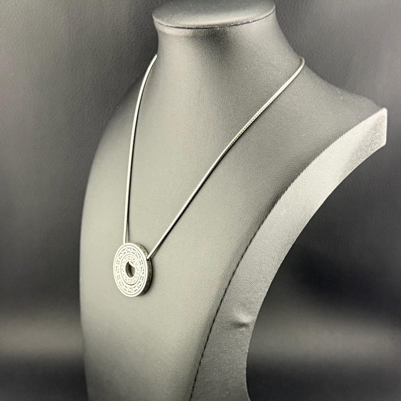 Puede incluir: Un collar con colgante circular de tono plateado que presenta un intrincado patr&oacute;n geom&eacute;trico repetitivo grabado en su superficie. El colgante est&aacute; suspendido de una cadena fina y lisa de tono plateado que descansa sobre el expositor.