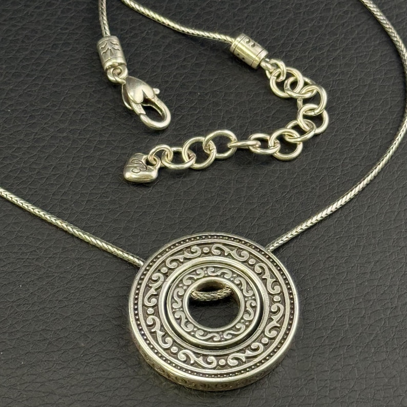 Puede incluir: Un collar de tono plateado con un colgante circular que presenta un intrincado patr&oacute;n de volutas en relieve. La cadena incluye un cierre de mosquet&oacute;n y una secci&oacute;n de extensi&oacute;n ajustable con un peque&ntilde;o dije en forma de coraz&oacute;n al final.