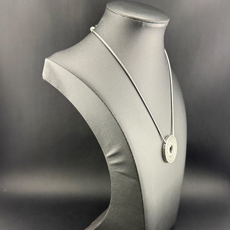 Puede incluir: Un collar de tono plateado que presenta una cadena fina y un colgante circular con un patr&oacute;n grabado detallado en su superficie. El colgante tiene un peque&ntilde;o recorte circular central y la pieza se exhibe sobre un busto de cuello negro.