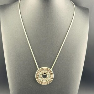 Puede incluir: Un collar de tono plateado con un colgante circular que presenta intrincados grabados de volutas y un recorte central. El colgante cuelga de una cadena fina y lisa de tono plateado que forma un bucle simple alrededor del expositor.