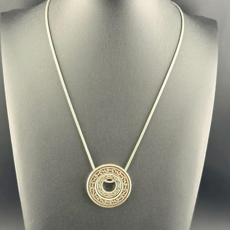 Puede incluir: Un collar de tono plateado con un colgante circular que presenta intrincados grabados de volutas y un recorte central. El colgante cuelga de una cadena fina y lisa de tono plateado que forma un bucle simple alrededor del expositor.