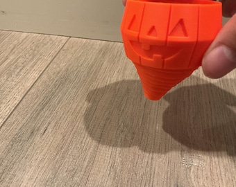 Puede incluir: Un objeto de color naranja brillante en forma de cono que presenta una cara de calabaza tallada con ojos triangulares y una amplia sonrisa dentada. La superficie tiene una textura acanalada que se estrecha hasta una punta, similar a una peonza.