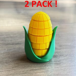 Puede incluir: Un objeto amarillo en forma de mazorca de maíz con una base verde. El objeto de plástico tiene un diseño detallado. El texto rojo en la parte superior dice "2 PACK!". Probablemente es un producto de cocina o relacionado con la comida.
