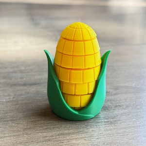 Puede incluir: Un objeto amarillo en forma de mazorca de maíz con un diseño segmentado, anidado dentro de hojas verdes. El maíz tiene una superficie texturizada, y las hojas proporcionan un borde verde contrastante. El objeto es probablemente un artículo decorativo o una pieza novedosa.