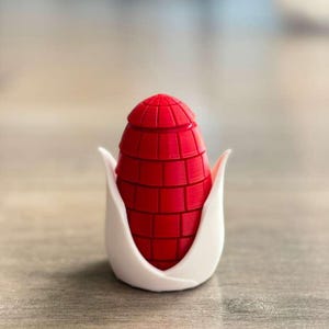 May include: A red, geometric-patterned object, resembling an ear of corn, is cradled in a white, petal-like holder. The object has a segmented design and a rounded top. The overall aesthetic is modern and artistic, with a focus on form and color.