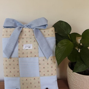 May include: A rectangular fabric sleeve featuring a patchwork design of cream-colored fabric with small red floral prints and light blue and white striped fabric. A light blue fabric bow is tied at the top. A white label reads Eternally June.
