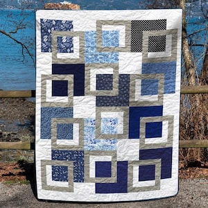 Puede incluir: Un edredón hecho a mano con un diseño geométrico. El edredón presenta cuadrados dentro de cuadrados en tonos de azul, gris y blanco. Algunos cuadrados tienen patrones florales, otros lunares. El edredón se exhibe al aire libre.