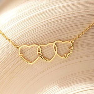 May include: A gold-colored necklace featuring three interconnected heart pendants. Each heart is inscribed with a name in a delicate script. The necklace is displayed on a neutral-toned surface, highlighting the jewelry's design.