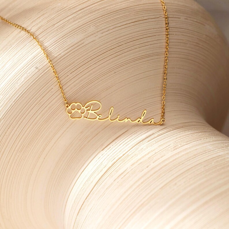 Puede incluir: Un delicado collar dorado con el nombre "Belinda" en cursiva. El dise&ntilde;o incluye un detalle de huella de pata al principio del nombre. El collar se muestra sobre un fondo neutro.