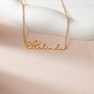 Puede incluir: Un collar dorado con el nombre "Belinda" en cursiva. Un dise&ntilde;o de huella de pata est&aacute; a la izquierda del nombre. El collar est&aacute; sobre una superficie rosa.