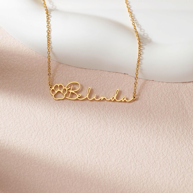 Puede incluir: Un collar dorado con el nombre "Belinda" en cursiva. Un dise&ntilde;o de huella de pata est&aacute; a la izquierda del nombre. El collar est&aacute; sobre una superficie rosa.