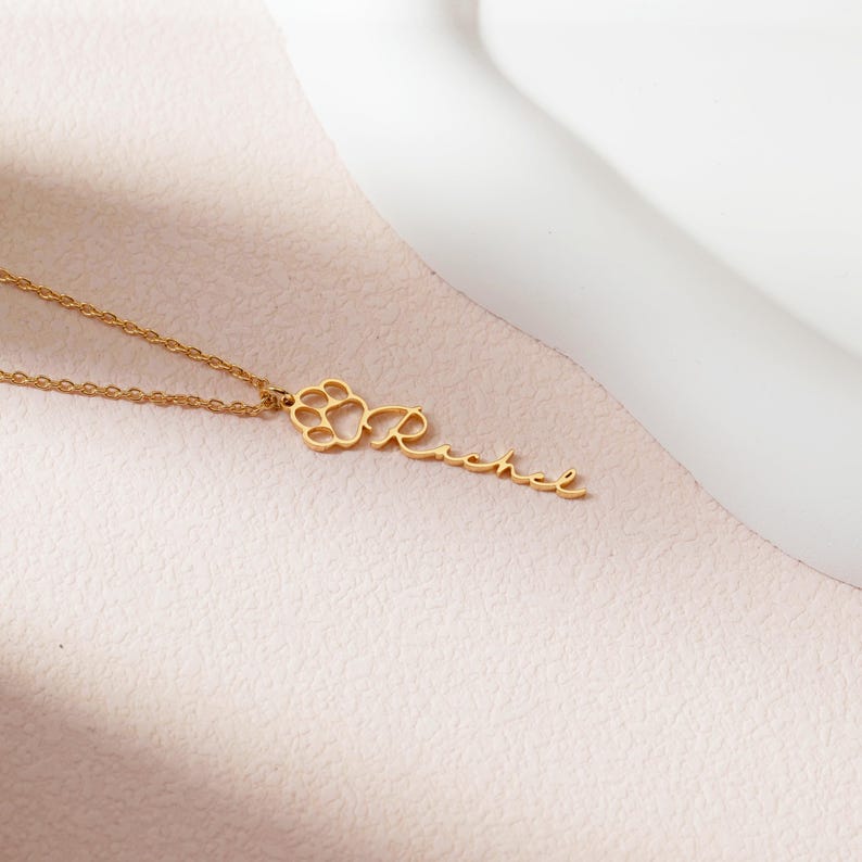 May include: A gold-colored necklace with a paw print design and the name "Rachel" in cursive script. The pendant is attached to a delicate chain. The necklace is displayed on a light pink surface.