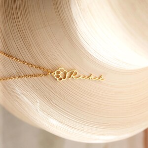 May include: A gold-colored necklace with a delicate chain. The pendant features a paw print design and the name "Rachel" in a cursive font. The necklace is displayed on a light-colored, textured surface.