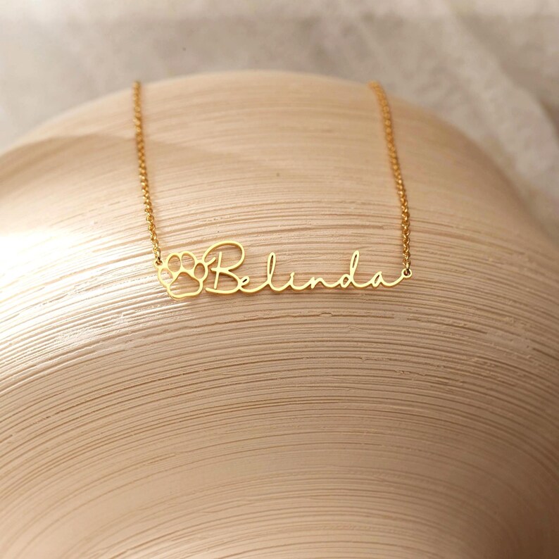 Puede incluir: Un collar dorado con el nombre "Belinda" en cursiva. La letra "B" est&aacute; dise&ntilde;ada con una huella de pata. El collar est&aacute; sobre una superficie de tonos neutros.
