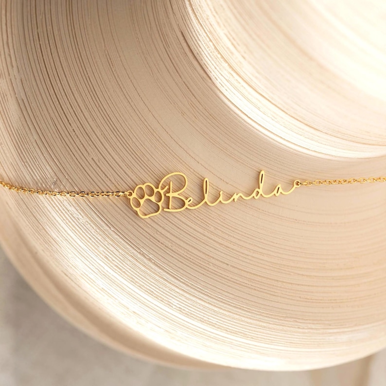 Puede incluir: Un collar dorado con el nombre "Belinda" en escritura cursiva. Un dise&ntilde;o de huella de pata est&aacute; a la izquierda del nombre. El collar est&aacute; sobre una superficie clara y texturizada.