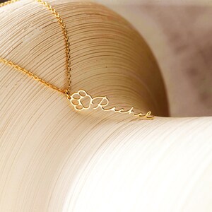 May include: A gold-colored necklace with a paw print charm and the name "Rachel" in a delicate script. The pendant is suspended from a fine chain, creating an elegant and personalized accessory. The necklace is displayed on a light-colored surface.