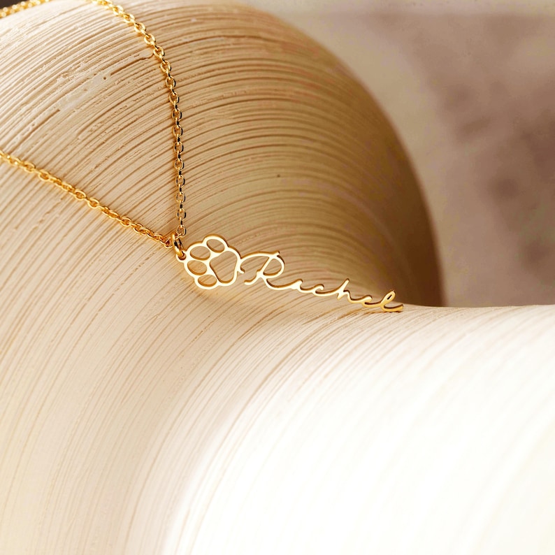 May include: A gold-colored necklace with a paw print charm and the name "Rachel" in a delicate script. The pendant is suspended from a fine chain, creating an elegant and personalized accessory. The necklace is displayed on a light-colored surface.