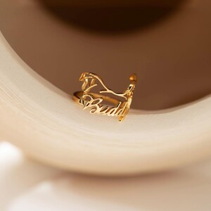 May include: A gold-toned metal ring featuring a cutout design of a dog silhouette and the name Buddy written in a cursive script font. The ring has an open band structure with a polished finish, showcasing the detailed lettering and animal shape.