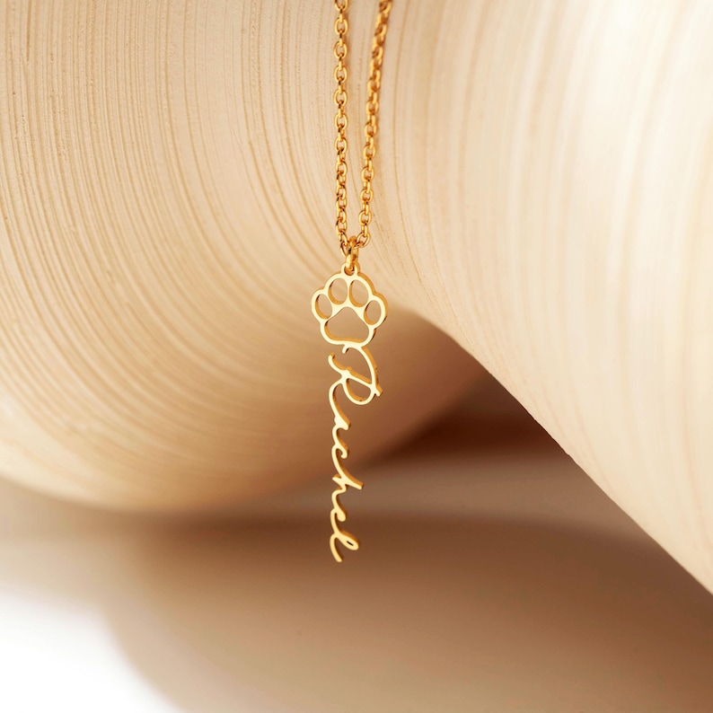 May include: Gold-colored necklace with a paw print charm and the name "Rachel" in a cursive font. The pendant hangs from a delicate chain. The background is a soft, neutral color.