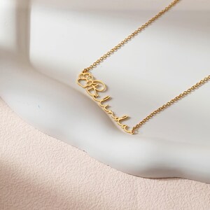 Puede incluir: Un collar dorado con el nombre "Linda" en cursiva. La letra "L" est&aacute; conectada a un dise&ntilde;o de huella de pata. La cadena es fina y el colgante est&aacute; centrado.