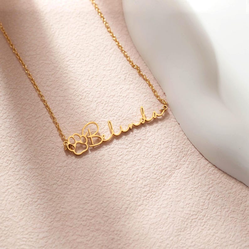Puede incluir: Un collar dorado con el nombre "Belinda" en cursiva. La letra "B" est&aacute; dise&ntilde;ada con una huella de pata. El collar se muestra sobre una superficie rosa claro.