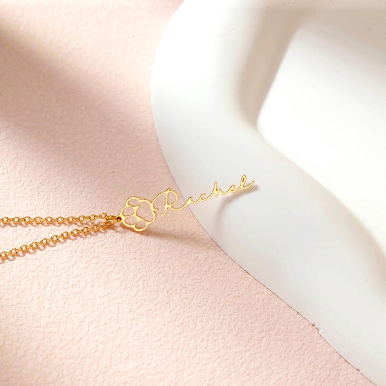 May include: A gold-colored necklace with a paw print charm and the name "Rachel" in cursive script. The paw print has a pink fill. The necklace is displayed on a light pink surface with a white curved object in the background.