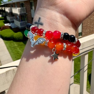 May include: Two beaded bracelets with a variety of colors, including red, orange, yellow, green, and black. One bracelet features a silver bat charm, and the other has a silver star charm. The bracelets are worn on a wrist.