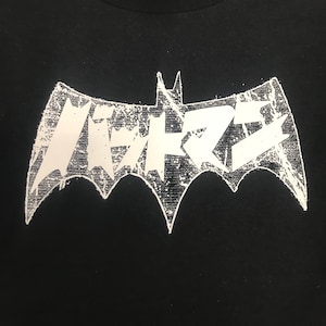 May include: Black t-shirt featuring a white, distressed bat-shaped logo with Japanese text. The logo has a textured, grunge-style appearance. The shirt is made of a dark fabric, and the logo is centered on the chest.