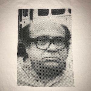 May include: White t-shirt featuring a black and white halftone portrait of a man wearing glasses. The image is a close-up of the man's face, with a slightly grumpy expression.