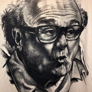 May include: Black and white graphic portrait of a man wearing glasses, with a surprised expression. The image is printed on a light-colored fabric, likely a t-shirt or similar apparel item. The style is reminiscent of a detailed sketch.