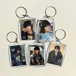 May include: Five clear acrylic keychains with silver key rings. Each keychain features a different image of a person. The keychains are rectangular with rounded corners. Each image has a star or heart design.