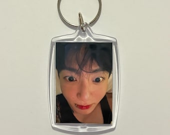May include: Clear rectangular keychain with a silver key ring. The keychain features a color photograph of a person with dark hair, a lip piercing, and red lipstick. The person is looking directly at the camera.