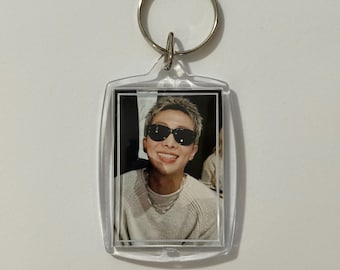 May include: A clear, rectangular keychain with a silver metal ring. The keychain features a smiling person wearing sunglasses. The person is wearing a light-colored sweater and a silver necklace. The background is blurred.