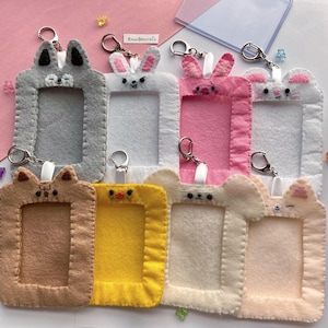 Könnte beinhalten: Acht Filz-Foto-Kartenhalter mit verschiedenen Tierdesigns. Jeder Halter hat ein transparentes Plastikfenster zum Einsetzen eines Fotos und einen Metallring zum Anbringen an einem Schlüsselbund. Die Designs umfassen einen grauen Hasen, einen weißen Hasen, einen rosa Hasen, einen weißen Hasen mit rosa Ohren, einen braunen Fuchs, ein gelbes Küken, einen weißen Bären und eine pfirsichfarbene Katze.