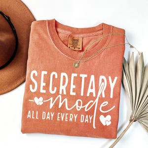 May include: A coral-colored Comfort Colors t-shirt with the words "SECRETARY mode ALL DAY EVERY DAY" in white script. A gold necklace with a pendant and a brown hat are also visible. The shirt is folded and displayed on a white surface.