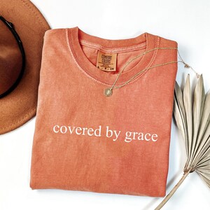 a t - shirt that says covered by grace next to a hat