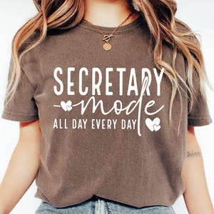 May include: A brown t-shirt with the text "SECRETARY mode ALL DAY EVERY DAY" in white lettering with two small white hearts.