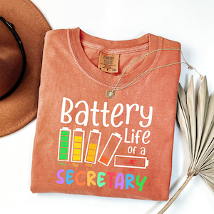 May include: A coral-colored Comfort Colors t-shirt with the text "Battery Life of a Secretary" in white and colorful letters. The design includes battery level indicators. A gold necklace and a brown hat are also visible.