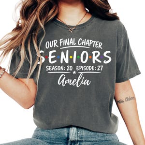 May include: A charcoal gray short-sleeve t-shirt featuring white text that reads OUR FINAL CHAPTER SENIORS SEASON: 20 EPISODE: 27 and the name Amelia. The word SENIORS includes small, colorful dots between the letters in red, blue, and orange.