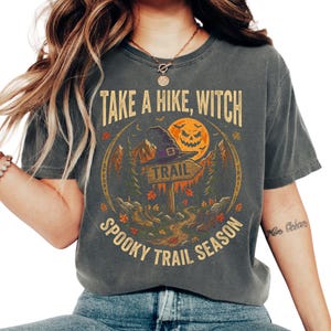 May include: A charcoal gray t-shirt featuring a graphic print with the text "TAKE A HIKE, WITCH" and "SPOOKY TRAIL SEASON". The design includes a witch hat on a trail sign, a jack-o'-lantern moon, mountains, trees, and hiking boots in orange and green.