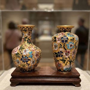 Może przedstawiać: Dwie wazy typu cloisonné ze złotym drutem i żywymi emaliowanymi wzorami kwiatowymi w odcieniach niebieskiego, różowego i zielonego. Wazy mają rozszerzane krawędzie i stoją obok siebie na prostokątnej, ciemnobrązowej drewnianej podstawce.