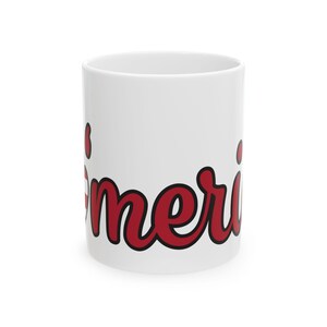 Puede incluir: Taza de cerámica blanca con la palabra "America" impresa en rojo cursivo en la parte delantera.